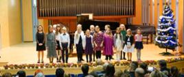 Zdjęcie przedstawia wszystkich uczestników koncertu stojących na scenie. Obok po prawej stronie choinka, za dziećmi fortepian i organy. Z przodu zdjęcia widownia . Przed dziećmi w ich nogach rozłożone są kwiatuszki. 