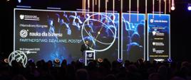 Zdjęcie od tyłu zaciemnionej sali, na oświetlonym podwyższeniu z przodu stoi minister Kulasek i mówi do mikrofonu, za nim na wielkim ekranie napis Partnerstwo - działanie - postęp.