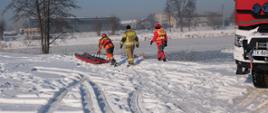 3 strażaków idzie z saniami lodowymi na lód, by móc ćwiczyć techniki ratowania. Obok zaparkowany samochód strażacki. W tle aura śnieżna. mroźna i staw skuty lodem.