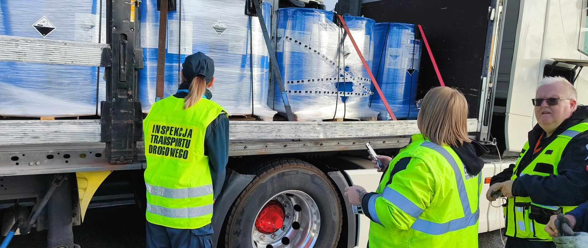 Trzy osoby w odblaskowych kamizelkach stoją obok naczepy ciężarówki z otwartą burtą. Jedna z osób, odwrócona tyłem, nosi żółtą kamizelkę z napisem INSPEKCJA TRANSPORTU DROGOWEGO. Pozostałe dwie osoby również mają na sobie odzież roboczą i przyglądają się przewożonemu ładunkowi. Na naczepie znajdują się beczki owinięte niebieską i przezroczystą folią stretch, zabezpieczone pasami transportowymi. Na pojemnikach widnieją symbole ostrzegawcze (piktogramy materiałów niebezpiecznych).
