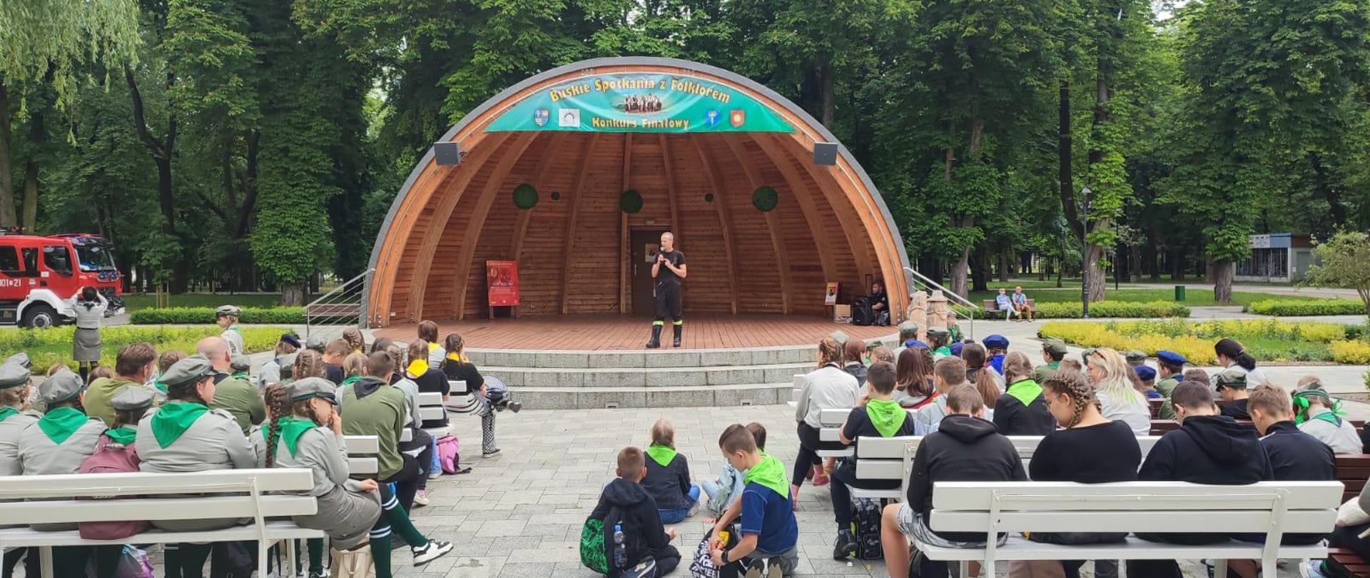 harcerze siedzą tyłem na białych ławkach, na muszli strażak opowiada. Po lewej stronie w tle samochód pożarniczy, w tle drzewa