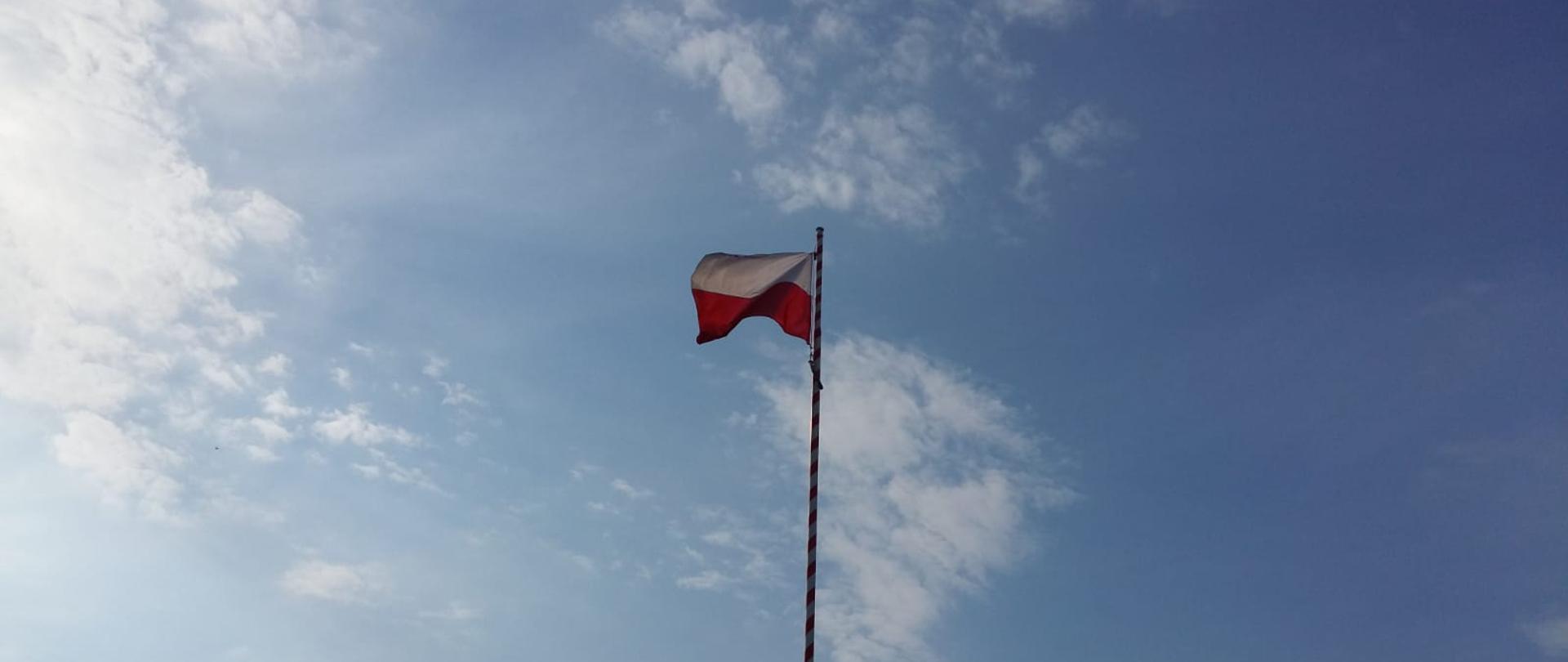 W centralnej części zdjęcia maszt z podniesioną flaga państwową, w tle błękitne niebo z niewielkimi chmurami.