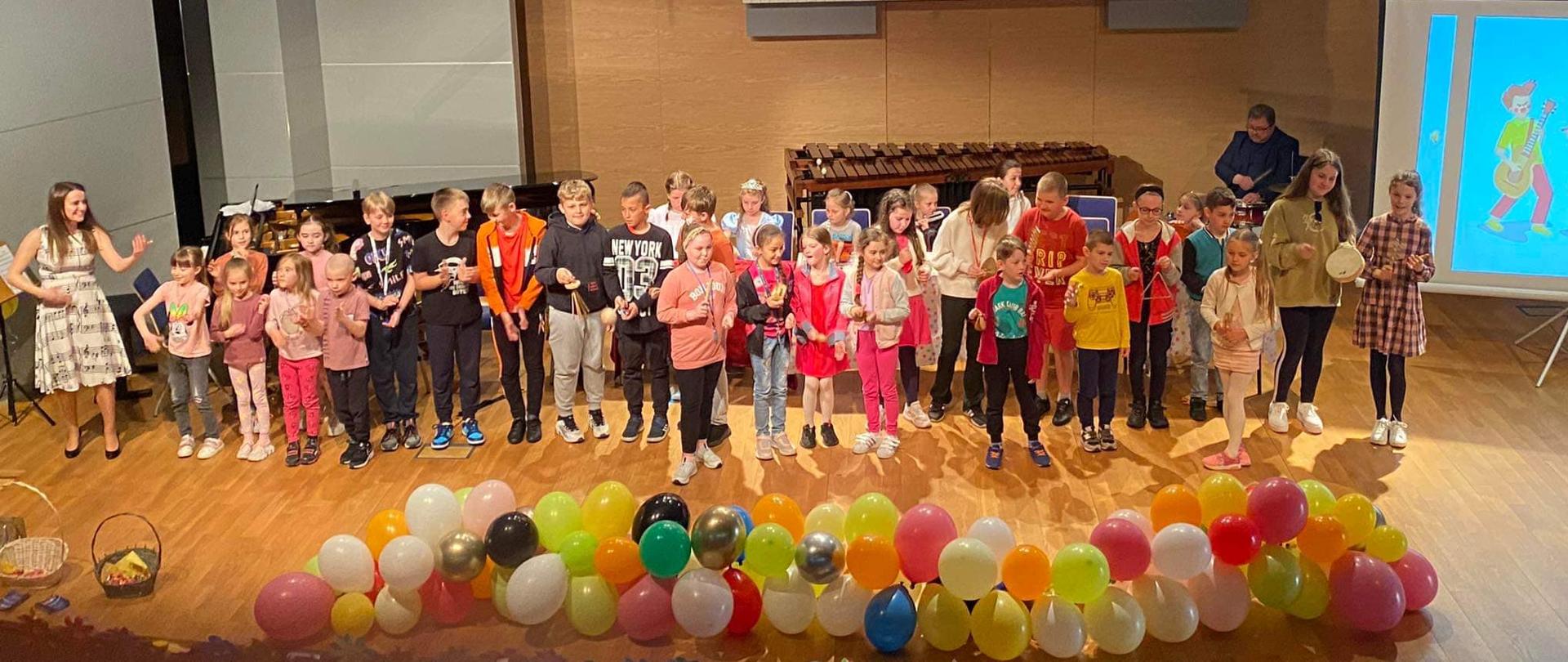 Zdjęcie sceny przedstawiające licznie zgromadzoną na niej występującą publiczność - przybyłe na koncert dzieci, przed nimi różne dekoracje między innymi w formie balonów. Po lewej stronie widoczny nauczyciel szkoły mgr Paulina Wilk ubrana w białą sukienkę z ikonografiką składającą się pięciolinii, oznaczeń muzycznych oraz nut
