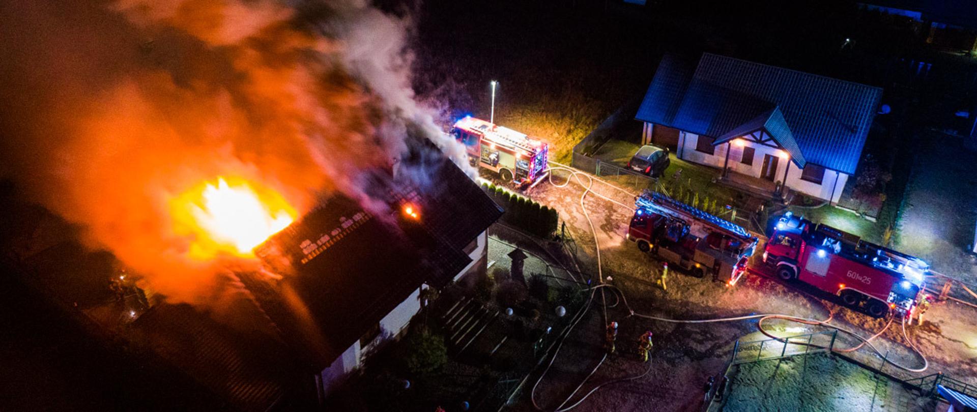 Na zdjęciu widok z lotu ptaka pojazdy roównlegle na jezdni dopalanego się domu, pożar wychodzi na dach budynku i strażacy gaszący palący się dom podający prądy gaśnicze 