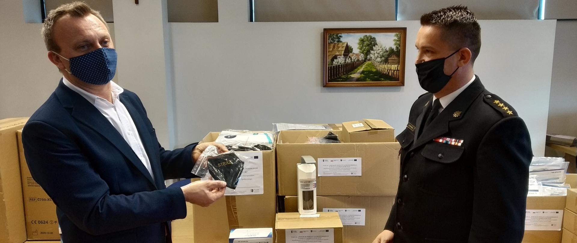 Zdjęcie przedstawia Starostę Marcina Piwnika, który prezentuje maseczkę z napisem straż, Po prawej komendant powiatowy PSP w mundurze wyjściowym patrzy w kierunku starosty. Za nimi na stole stoją pudełka ze środkami ochrony indywidualnej