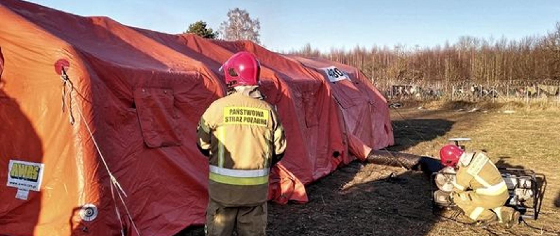 Namiot PSP koloru pomarańczowego rozstawiony w ramach wsparcia logistycznego na granicy, jeden strażak stoi obok namiotu, drugi w pozycji klęczącej działa przy nagrzewnicy do namiotu, w tle granica i las