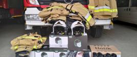 Zdjęcie przedstawia nowozakupiony sprzęt umieszczony w garażu na tle samochodów ratowniczo-gaśniczych. Strażacy zakupili ubrania specjalne koloru piaskowego, białe hełmy, granatowe rękawice, czarne buty.