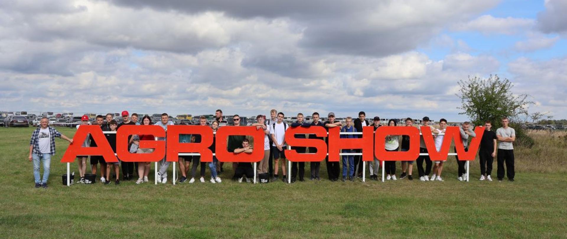 Grupa uczniów i nauczyciela zgromadzona za dużym czerwonym napisem AGRO SHOW