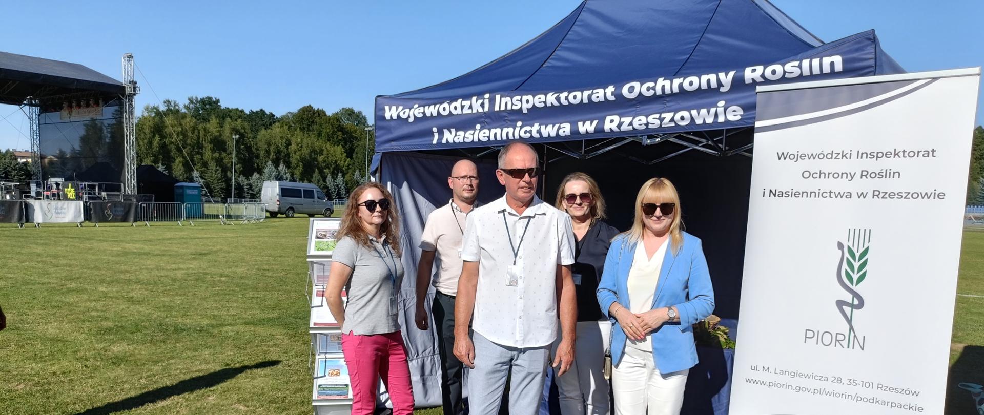 Grupowe zdjęcie pięciorga pracowników Wojewódzkiego Inspektoratu Ochrony Roślin i Nasiennictwa w Rzeszowie przed granatowym namiotem informacyjnym. Osoby stoją na trawiastym boisku i patrzą w stronę aparatu. W centrum znajduje się mężczyzna w jasnej koszuli i szarych spodniach, obok niego stoją pozostali członkowie zespołu, ubrani między innymi w marynarkę, koszulkę polo oraz jasną marynarkę i białe spodnie. Kilka osób ma okulary przeciwsłoneczne i identyfikatory na smyczach. Po prawej stronie znajduje się duży roll-up z nazwą Wojewódzkiego Inspektoratu Ochrony Roślin i Nasiennictwa w Rzeszowie oraz logo PIORiN. W tle widać scenę plenerową, drzewa, ogrodzenie oraz inne elementy wydarzenia. Jest słoneczny, bezchmurny dzień.