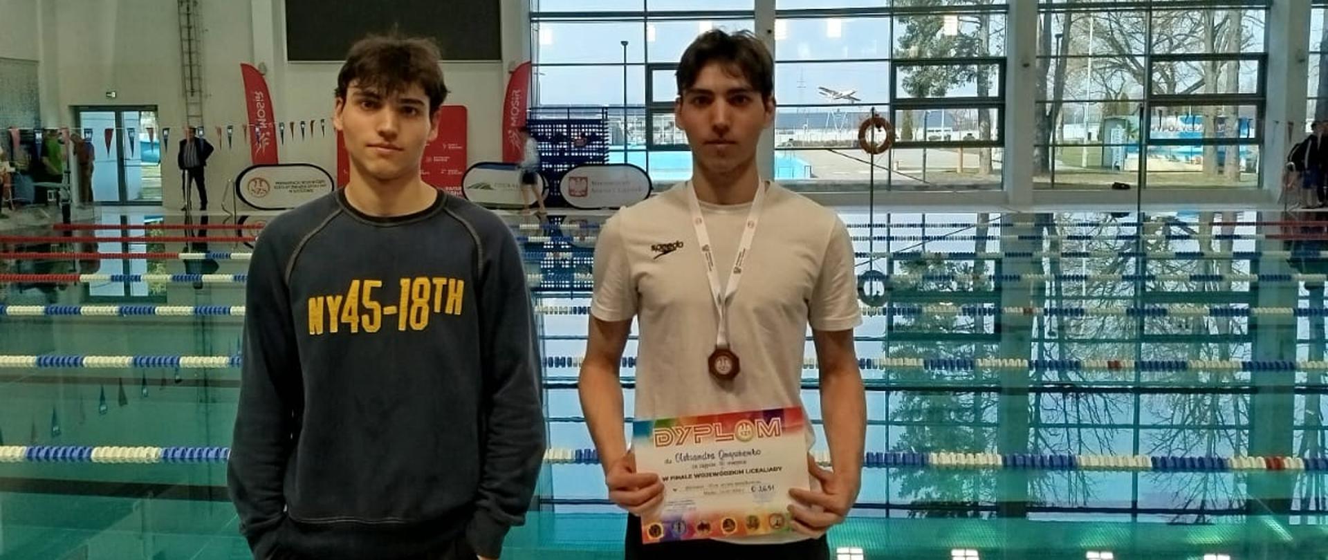Na zdjęciu znajduje się dwóch uczniów ZSCKR na tle basenu, jeden z uczniów trzyma dyplom