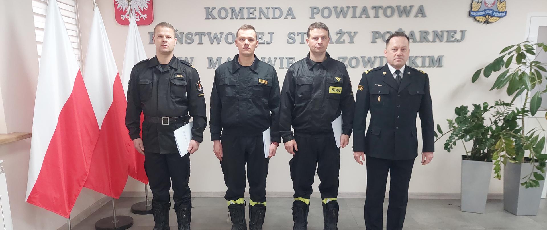 Trzech wyróżnionych funkcjonariuszy oraz Komendant Powiatowy PSP stoją w szeregu na tle ściany z napisem Komenda Powiatowa Państwowej Straży Pożarnej w Makowie Mazowieckim oraz Godło Polski i logiem PSP. Po ich prawej stronie znajdują się 3 Polskie Flagi na stojakach.