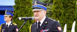 Kujawsko-pomorski Komendant Wojewódzki Państwowej Straży Pożarnej w granatowym mundurze galowym stoi przy mikrofonie i mównicy podczas oficjalnych obchodów dnia strażaka w Rypinie. W tle widoczne są biało-czerwone flagi oraz ozdobne drzewa otaczające plac jednostki.