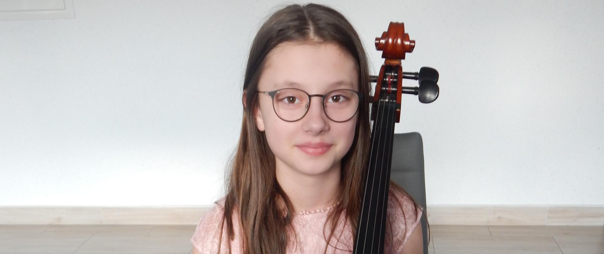 Dziewczynka w okularach siedzi przy wiolonczeli, w ręku trzyma smyczek. Ma na sobie sukienkę w kolorze jasnego różu. W tle biała ściana a na podłodze granatowa wykładzina. 