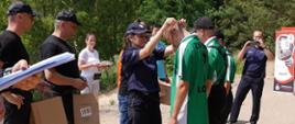 Zdjęcie przedstawia oficera prasowego komendanta powiatowego policji w Łowiczu komisarz Urszulę Szymczak, która wręcza pamiątkowe medale uczestnikom zawodów, za jej placami stoją komendant powiatowy PSP w Łowiczu mł. bryg. Arkadiusz Makowski oraz zastępca komendanta powiatowego PSP w Łowiczu mł. bryg Rafał Garbarz, którzy trzymają w rękach papierowe torby z nagrodami dla zawodników, w tle policjant robi zdjęcie na tle pobliskiego lasu, gratulację składa zawodnikom przedstawiciel Wojewódzkiej Stacji Ratownictwa Medycznego w Łodzi.