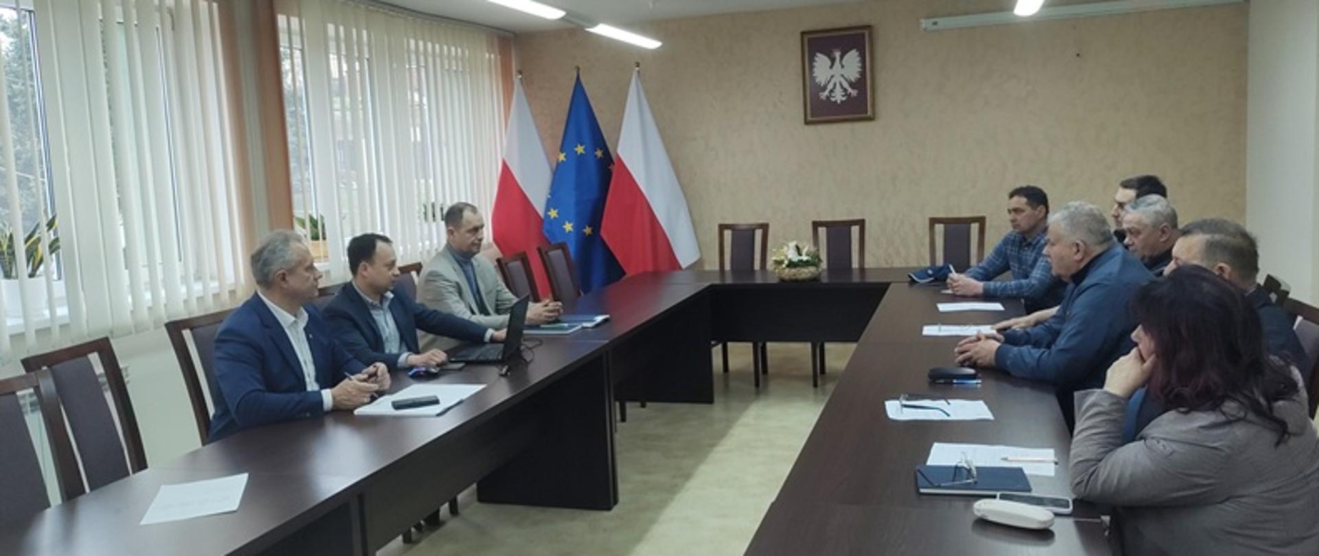 Grupa osób siedząca w jasnym pomieszczeniu. W tle widać flagi.