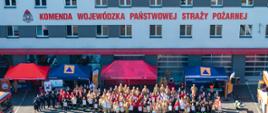 Szeroki widok z góry na elewację budynku Komendy Wojewódzkiej PSP w Opolu. Biały budynek z czerwonymi akcentami i rzędami okien. Na górze duży czerwony napis: „KOMENDA WOJEWÓDZKA PAŃSTWOWEJ STRAŻY POŻARNEJ”. Przed budynkiem, na placu, stoi bardzo duża grupa ludzi – kilkaset osób – ustawionych do wspólnego zdjęcia. Wszyscy w różnych mundurach strażackich (dorośli i dzieci, w tym w mundurach młodzieżowych). Przed grupą, pośrodku, widać dwa czerwone namioty. Lewy namiot z numerem alarmowym 112, prawy z napisem na niebieskim banerze. Plac jest pusty przed grupą, widać tylko cienie. W tle u góry, na dachu, widać instalację fotowoltaiczną."