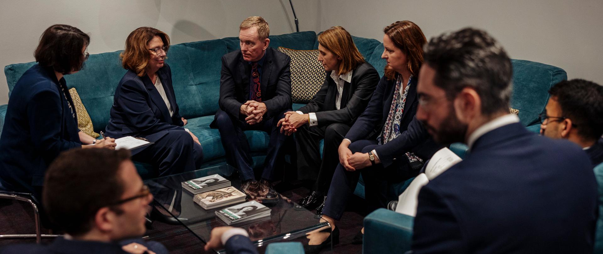 Osiem osób ubranych w garnitury siedzi na niebieskich kanapach i fotelach. Na środku jest szklana ława, na której są książki. 