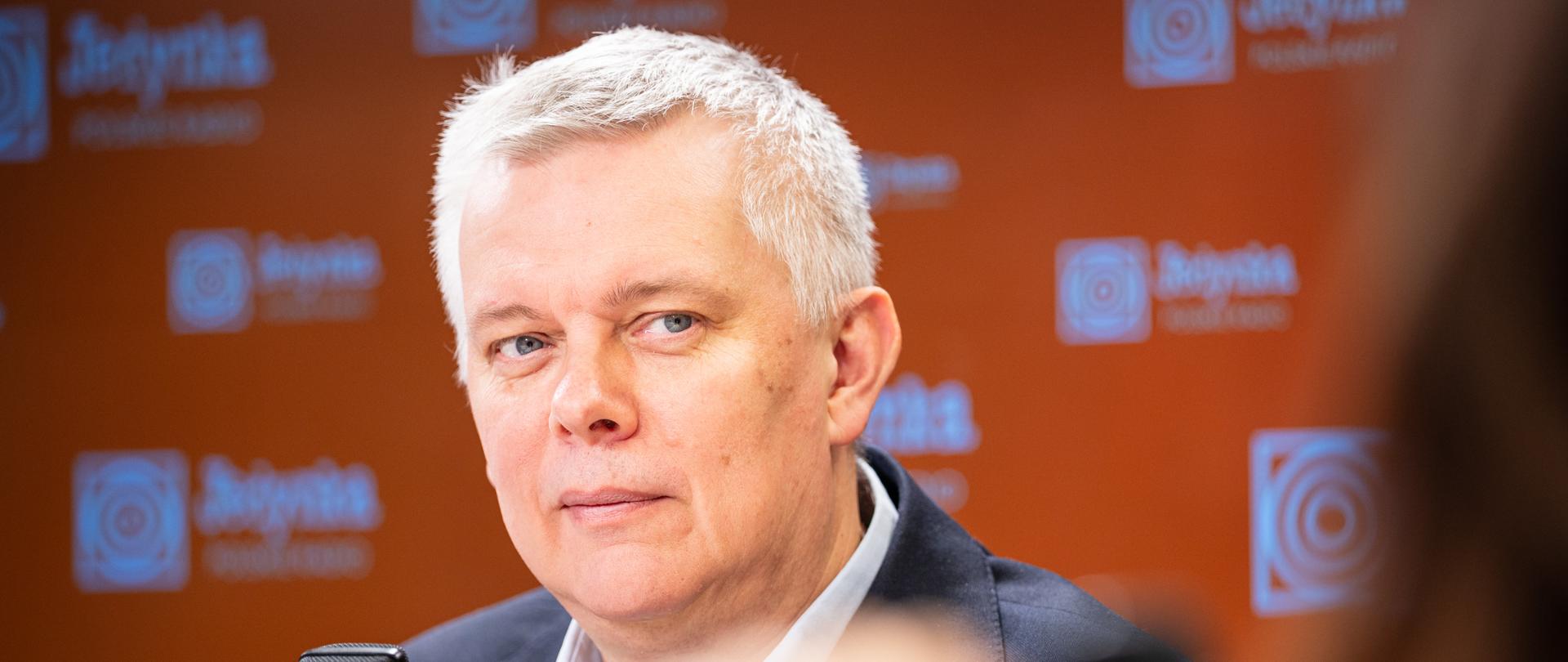 Na zdjęciu minister Tomasz Siemoniak w studiu radiowym. W lewym dolnym rogu zdjęcia widać mikrofon. W tle pomarańczowa ścianka z logo radiowej Jedynki.