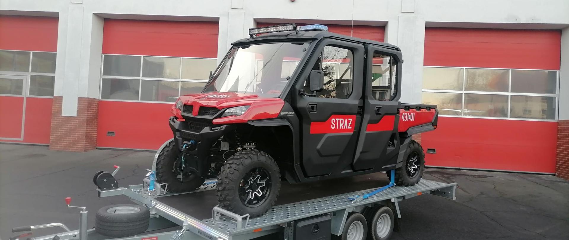 Zdjęcie przedstawia quada typu UTV znajdującego się na lawecie. Zdjęcie zrobione na placu przed komendą.