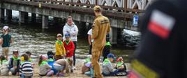 Strażak w umundurowaniu koloru piaskowego tłumaczy dzieciom znajdującym się na plaży zasady bezpiecznego przebywania na plaży. W tle widoczne jest jezioro oraz molo.