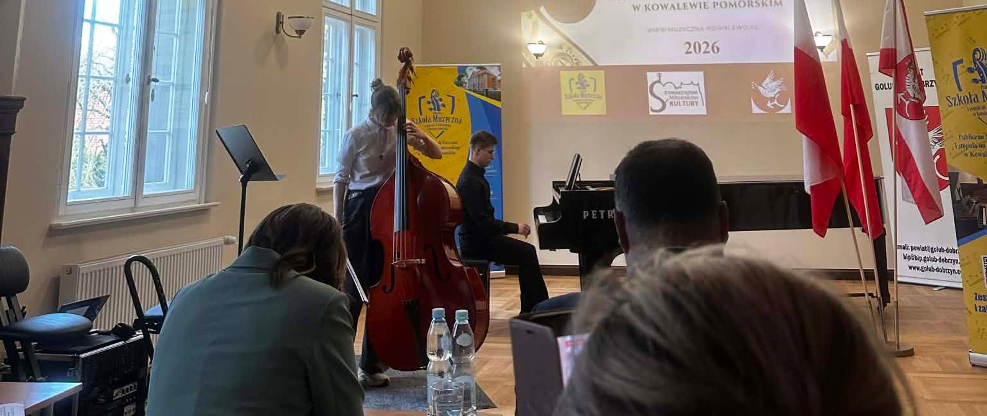 djęcie przedstawia występ muzyczny podczas XII Ogólnopolskiego Konkursu Wiolonczelowo-Kontrabasowego w Kowalewie Pomorskim. Na scenie widoczna jest młoda artystka grająca na kontrabasie oraz akompaniujący jej pianista przy czarnym fortepianie marki Petrof. W tle znajduje się ekran z logotypem konkursu oraz datą 2026, a obok stoją flagi Polski. Nad sceną zamontowana jest profesjonalna rampa z oświetleniem scenicznym i nagłośnieniem. Na pierwszym planie widoczne są plecy osób siedzących na widowni oraz stół sędziowski z butelkami wody i mikrofonem.