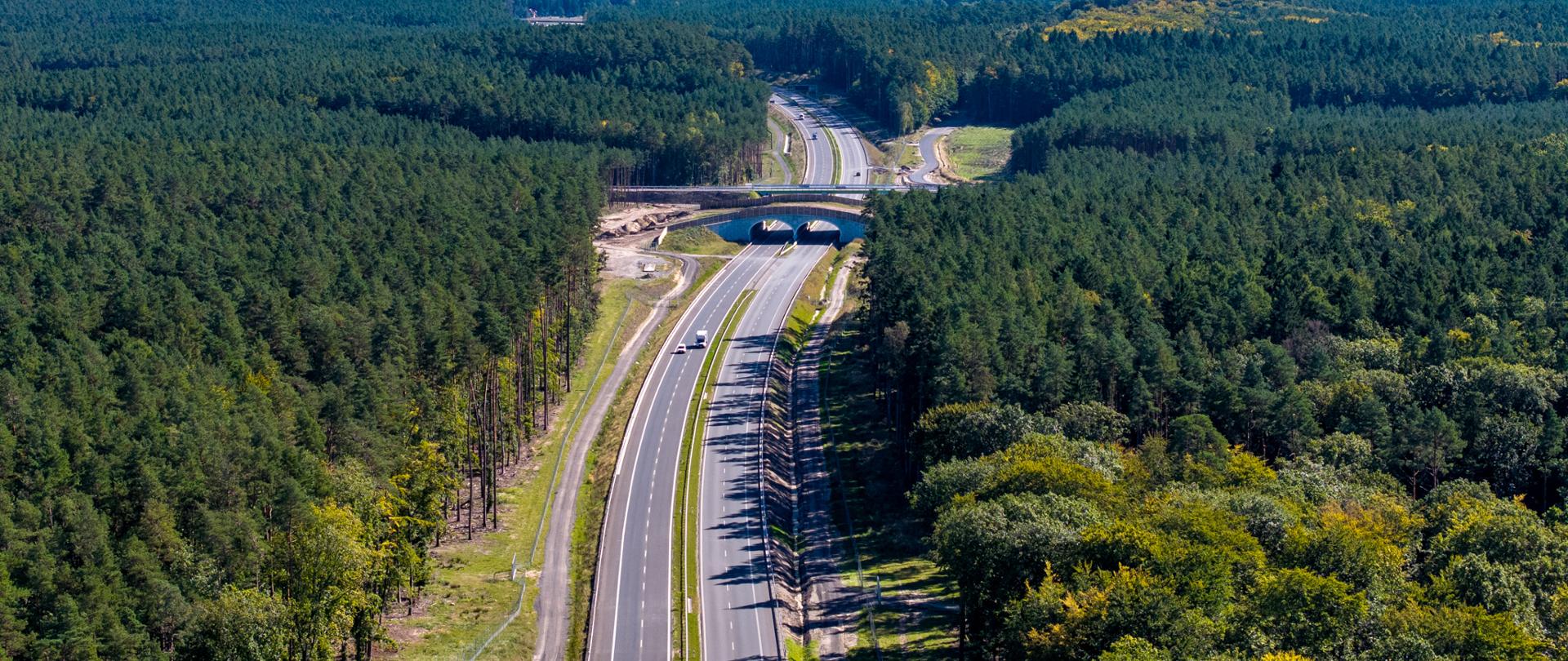 Zdjęcie lotnicze przedstawiające dwupasmową drogę przebiegającą przez gęsty, zielony las. Na środku kadru widać wiadukt nad trasą, a dalej rozciąga się szeroki, leśny krajobraz aż po horyzont. Na drodze jedzie kilka samochodów. 