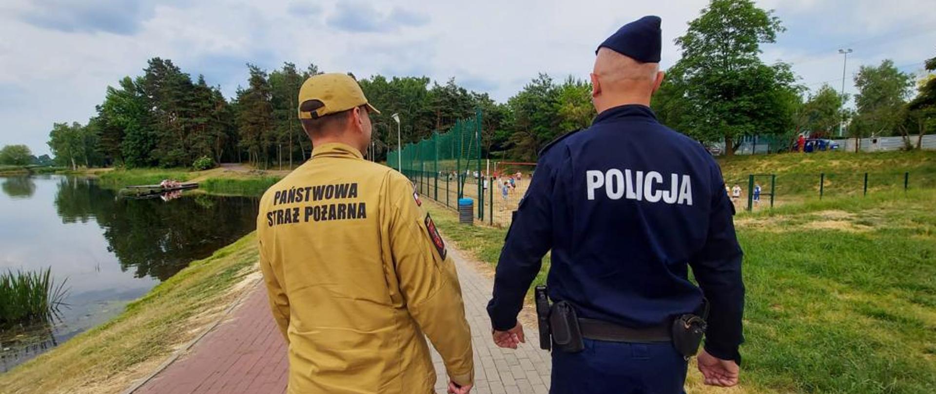 Dwóch mężczyzn, funkcjonariuszy idzie deptakiem nad zbiornikiem wodnym. Mężczyzna po lewej stronie jest ubrany w mundur w kolorze piaskowym z napisem na plecach Państwowa Straż Pożarna, a drugi mężczyzna ten po prawej stronie ubrany jest w granatowy mundur z napisem na plecach Policja.