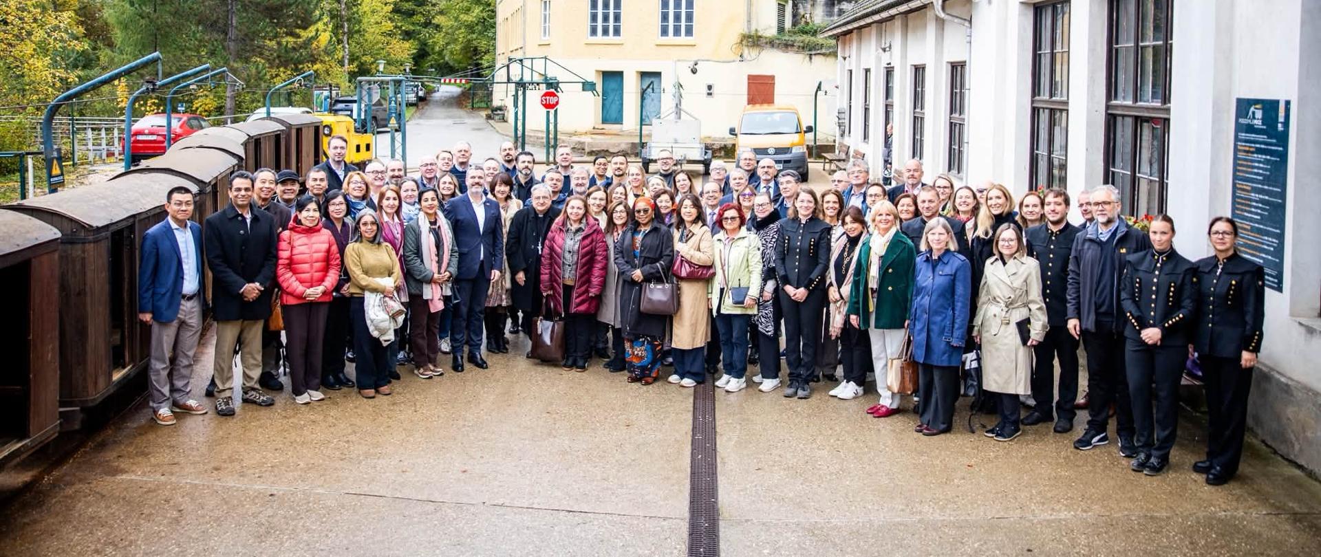 Večja skupina več deset ljudi pozira za skupinsko fotografijo na prostem pred stavbami nekdanjega rudnika v Mežici. Na levi so manjši rudniški vozički na tirih, na desni bela stavba z velikimi okni. V ozadju so druge stavbe in gozdnata pobočja v jesenskih barvah.