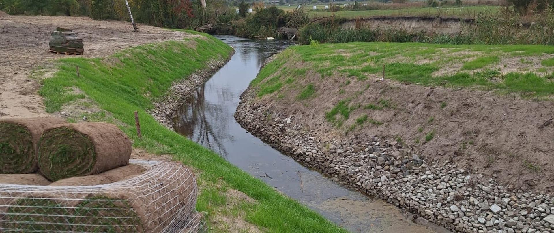Przepławka w Pakości przypomina naturalne koryto małej rzeczki. Jest prawie gotowa. Brzegi umocnione kamieniami. Po lewej stronie stoi paleta, a na niej zwinięte rolki trawy. 