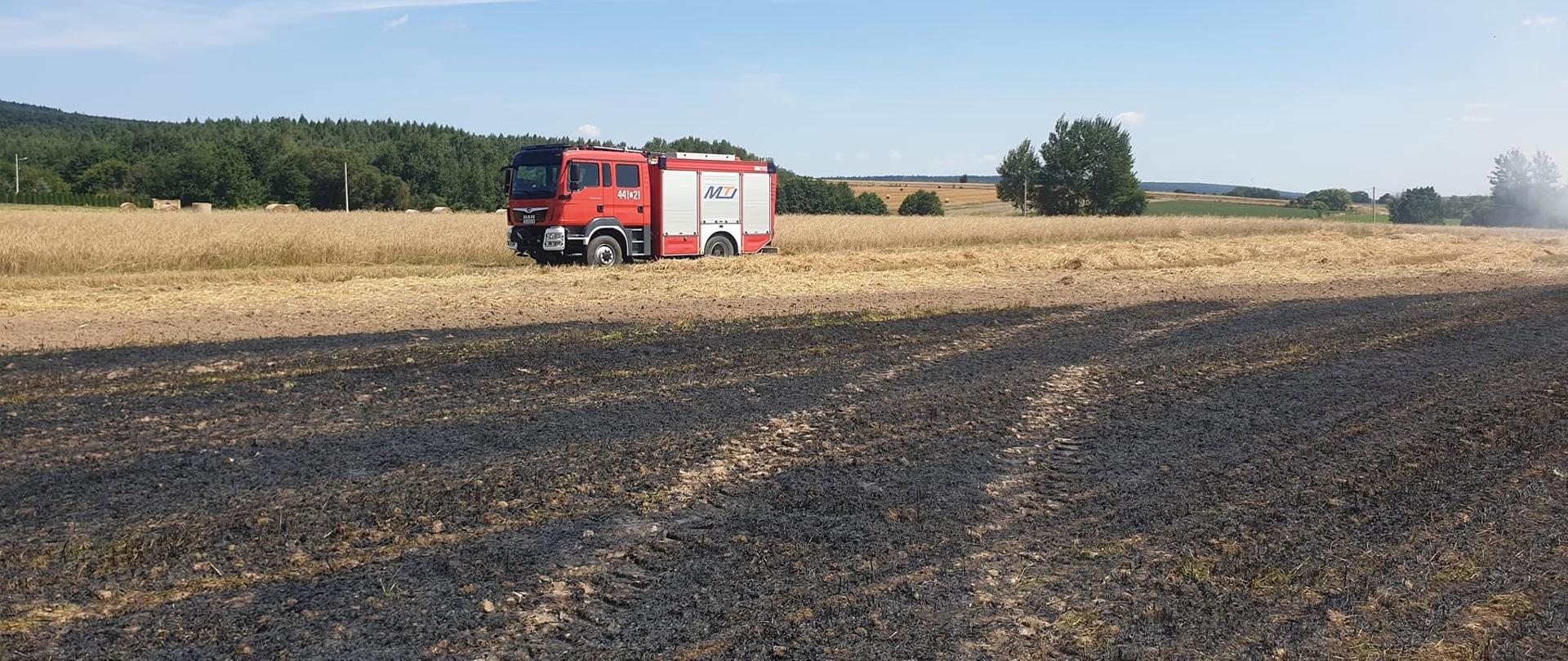 Samochód ratowniczo-gaśniczy stoi na skraju pola zboża obok pogorzeliska po pożarze ścierniska. W tle rozciągają się pola uprawne i las.