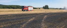 Samochód ratowniczo-gaśniczy stoi na skraju pola zboża obok pogorzeliska po pożarze ścierniska. W tle rozciągają się pola uprawne i las.