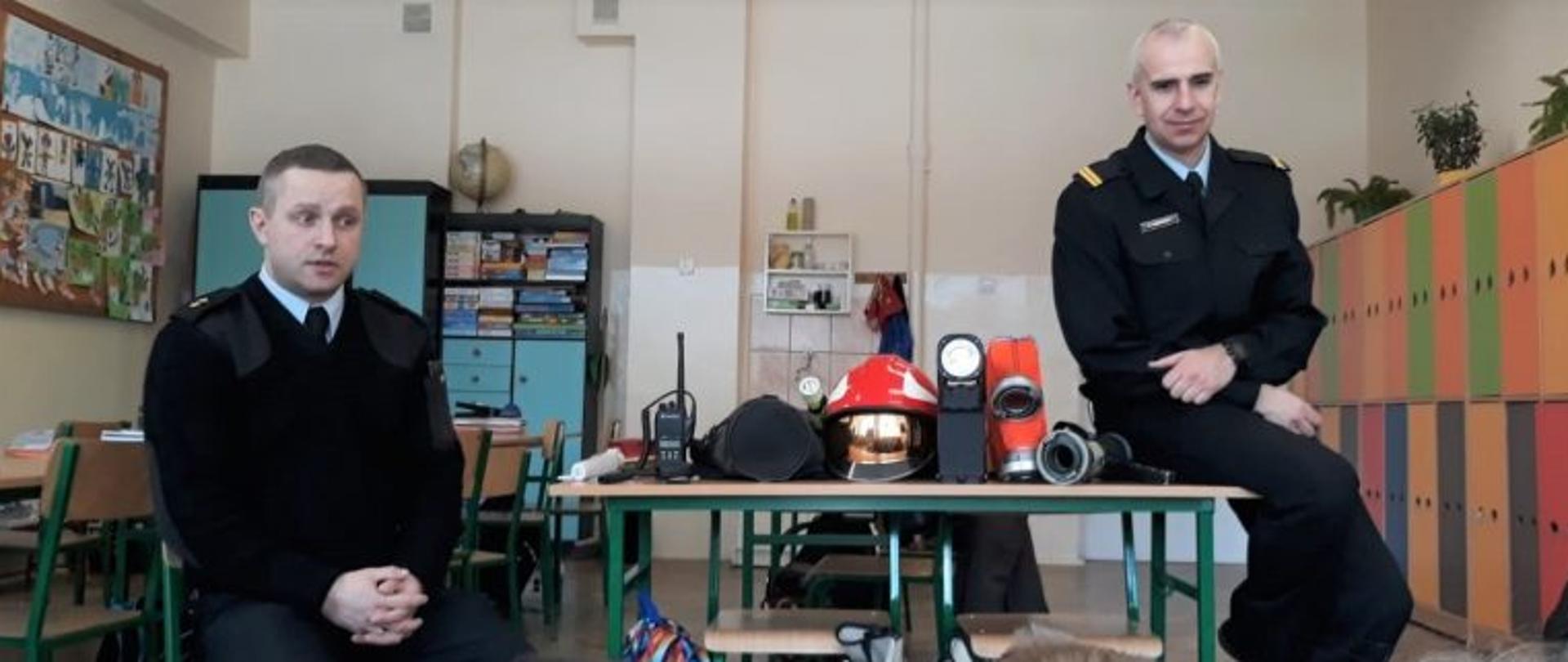 Strażacy komendy powiatowej PSP w umundurowaniu służbowym prezentują podstawowy sprzęt z wyposażenia strażaka między innymi obuwie, hełm, wąż, prądownice i sprzęt łączności. Poniżej widać siedzące i słuchające dzieci. Wydarzenie ma miejsce w klasie szkolnej.