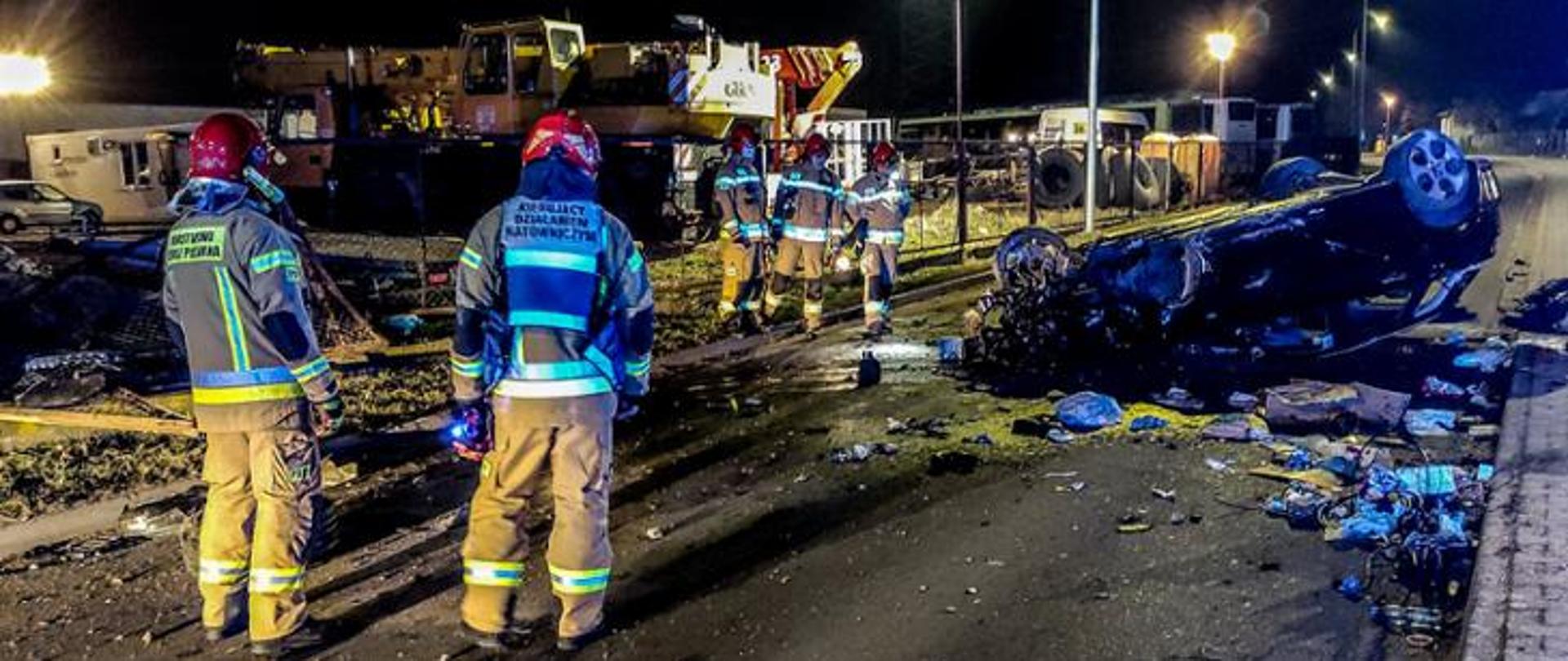 leżacy na dachu uszkodzony samochów, w pobliżu strażacy podczas działań ratowniczych