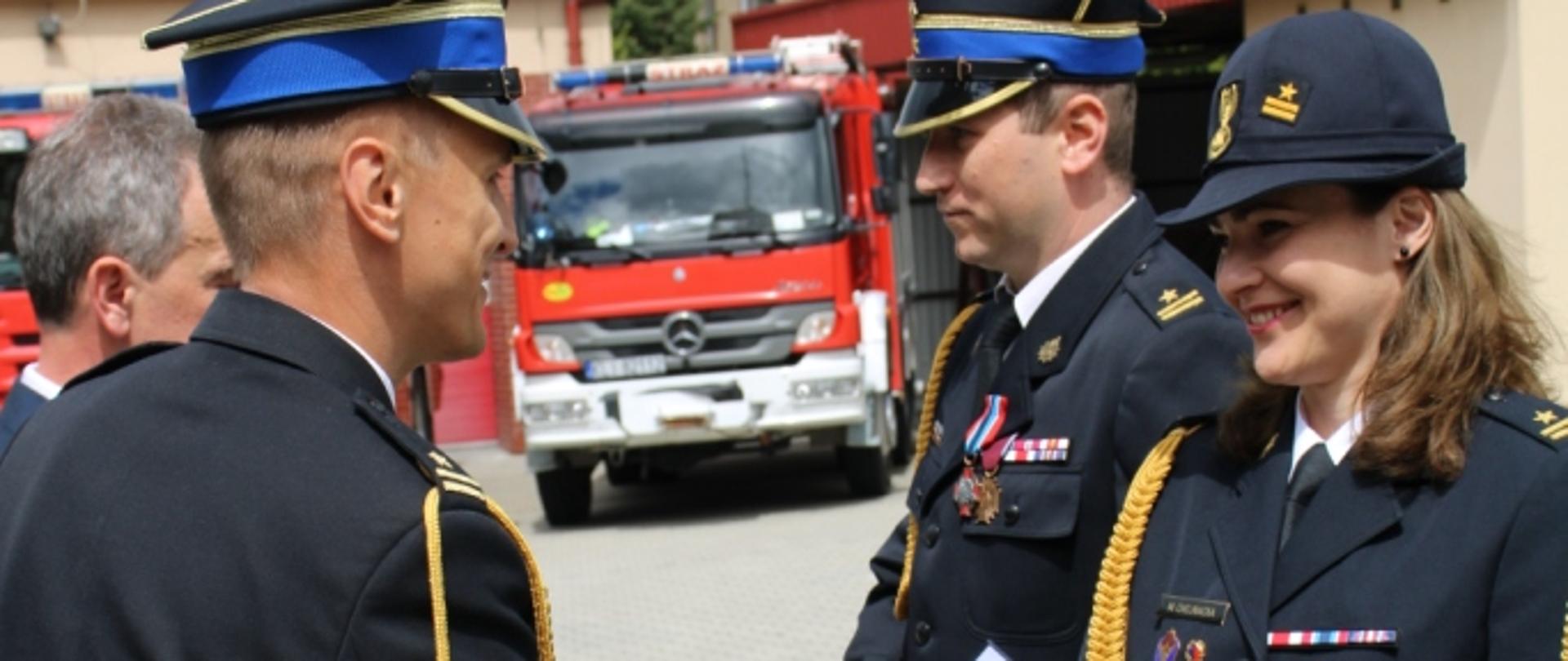 Na zdjęciu Zastępca Małopolskiego Komendanta Wojewódzkiego PSP w Krakowie mł. bryg. Przemysław Brzęczek wręcza akt nadania wyższego stopnia służbowego dla funkcjonariuszy KP PSP Limanowa st. kpt. Monika Chojnacka i st. kpt. Tomasz Kaptur. W tle pojazdy pożarnicze i obiekty KP PSP Limanowa.