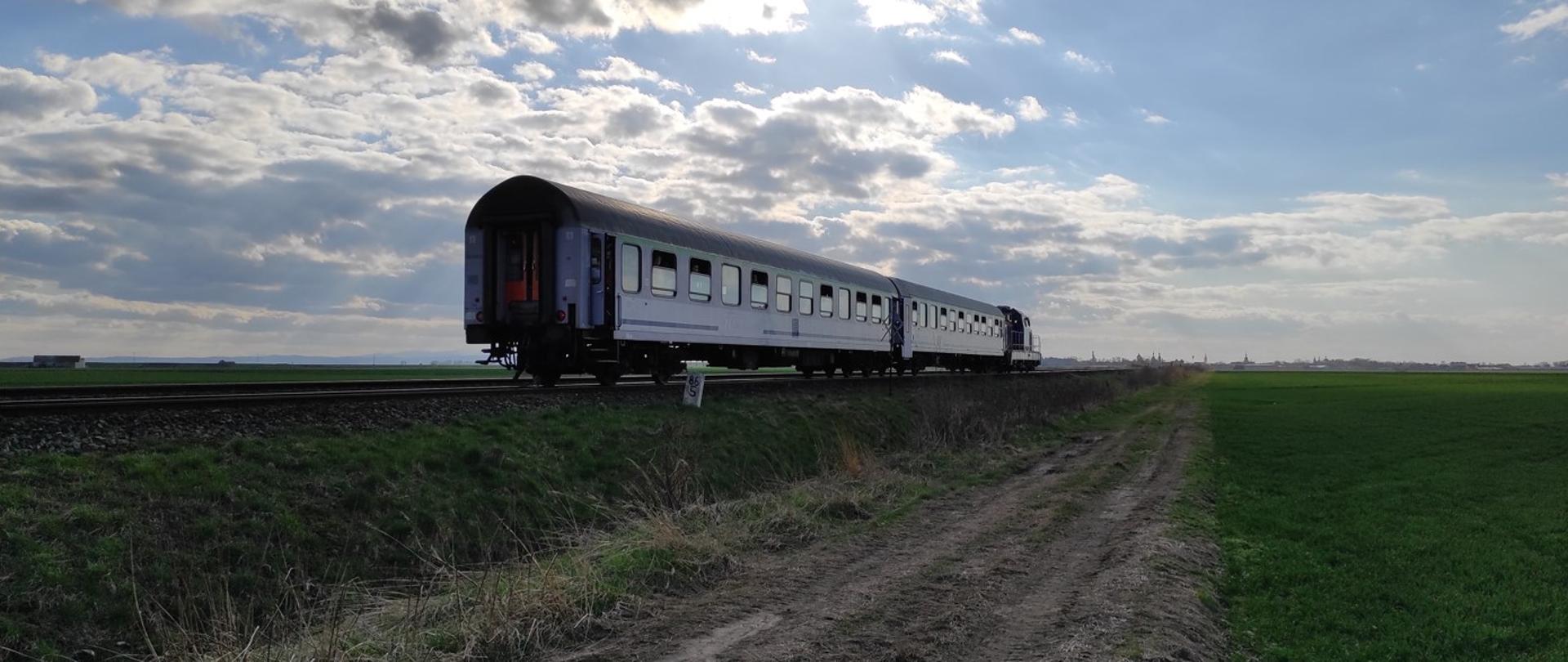 Zdjęcie przedstawia miejsce zdarzenia po wypadku na przejeździe kolejowym. Na zdjęciu widoczny pociąg osobowy składający się z lokomotywy i dwóch wagonów osobowych
