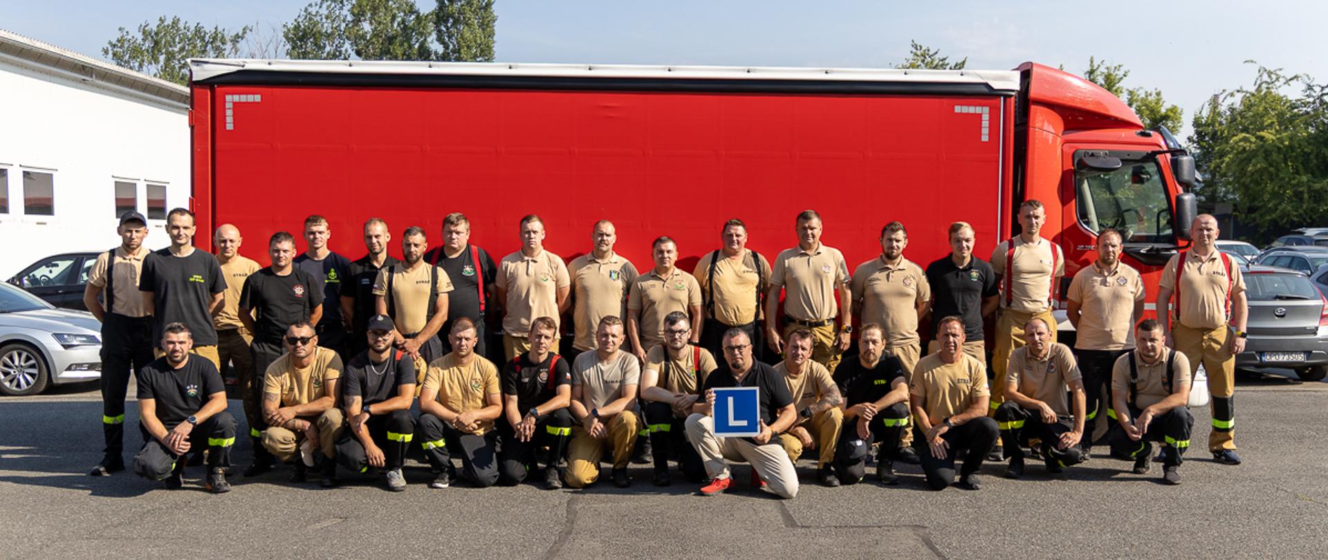 Około dwudziestopięcioosobowa grupa strażaków ratowników OSP stoi na placu przed dużym czerwonym samochodem ciężarowym wykorzystywanym w szkoleniu. Uczestnicy mają na sobie beżowe lub czarne koszulki i jasne spodnie, część z elementami odblaskowymi. Wszyscy pozują do wspólnego zdjęcia, ustawieni w kilku rzędach. Jedna osoba w pierwszym rzędzie trzyma niebieską tabliczkę z białą literą „L”, oznaczającą pojazd szkoleniowy. W tle widać zaparkowane samochody i budynki. Zdjęcie dokumentuje rozpoczęcie drugiego szkolenia przygotowującego do egzaminu na kierowanie pojazdem powyżej 3,5 t dla strażaków OSP.