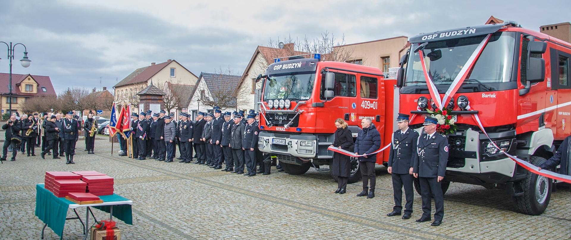Zdjęcie przedstawia strażaków w trakcie uroczystego przekazania nowego pojazdu gaśniczego dla OSP Budzyń.
W tle rynek budzyński i budynki.
