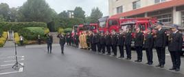 Po prawej stronie – budynek JRG przed którym ustawiono pojazdy pożarnicze. Na ich tle od lewej do prawej ustawiony pododdział pożarniczy – większość strażaków w mundurach galowych. Część w piaskowych mundurach służbowych. Na środku widać przechodzących wzdłuż pododdziałów Zastępcę Komendanta Wojewódzkiego w asyście dowódcy uroczystości dokonującego przeglądu pododdziału.