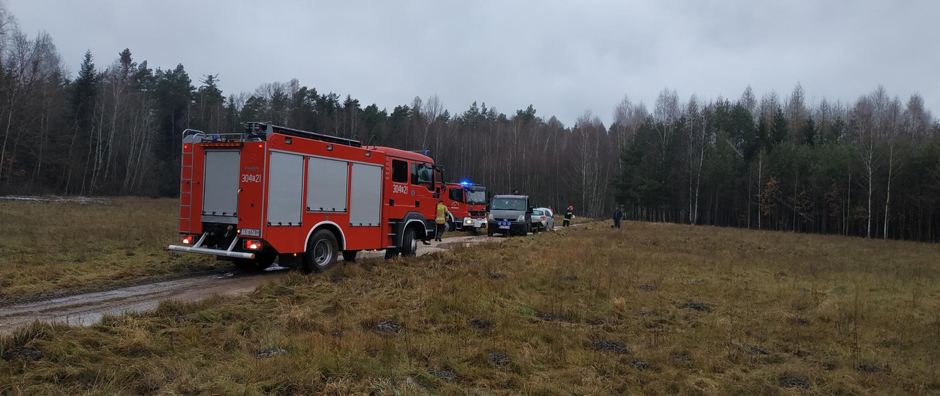 Zdjęcie przedstawia samochód pożarniczy na środku wąskiej drogi pod lasem. W oddali widać samochód osobowy oraz drugi samochód straży pożarnej. Nie widać ognia ani dymu. 