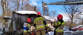 Widok ogólny – na pierwszym planie trzech strażaków widocznych od tyłu, w tle budynek mieszkalny z zawalonym dachem, po prawej stronie wysunięta drabina mechaniczna nad dachem obiektu.
