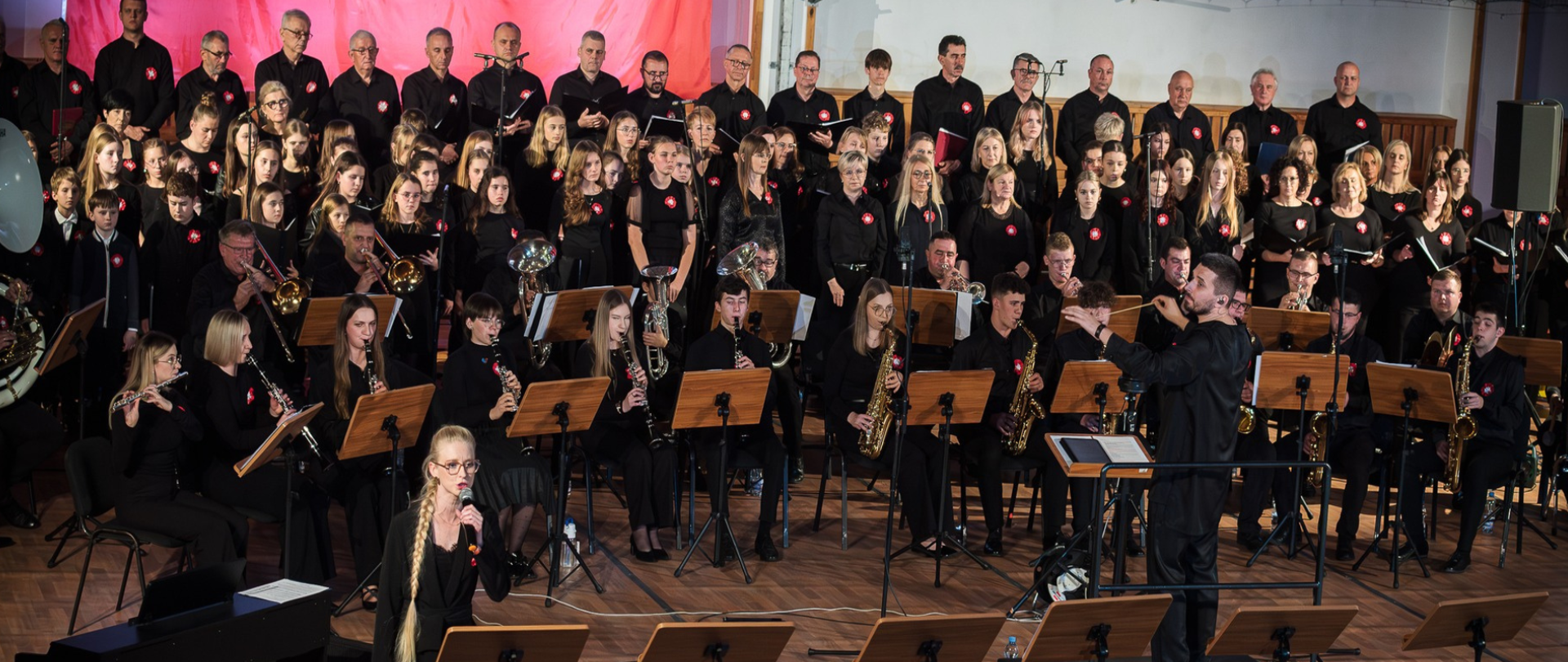 Duża grupa wykonawców podczas koncertu patriotycznego. W centrum kadru widoczna jest orkiestra dęta i instrumentaliści, w tym saksofoniści, fleciści oraz trębacze, a także dyrygent stojący tyłem do widza. Przed nimi, na pierwszym planie, młoda dziewczyna w okularach śpiewa do mikrofonu. Za orkiestrą, na podwyższeniu, ustawiony jest duży chór złożony z dorosłych i młodzieży. Wszyscy wykonawcy są ubrani na czarno i mają przypięte biało-czerwone kotyliony lub symboliczne znaczki w kształcie Polski. Tło stanowi częściowo czerwono-biała dekoracja.