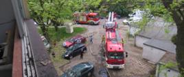 Widok z okna w którym miał miejsce pożar, przed blokiem na parkingu znajdują się trzy samochody pożarnicze, rozwinięte są linie gaśnicze, pracują strażacy