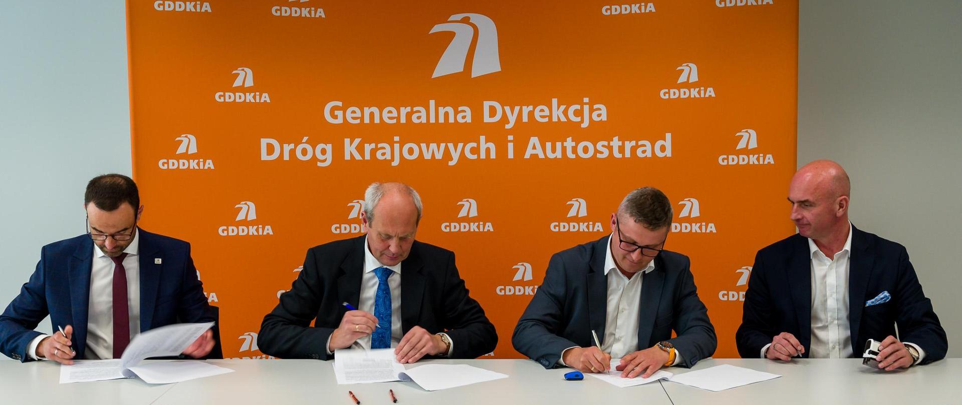 Moment podpisania jednego z 18 aneksów waloryzacyjnych. 4 elegancko ubranych mężczyzn siedzących obok siebie podpisuje dokumenty. W tle pomarańczowa ścianka z logotypami i nazwą GDDKiA.
