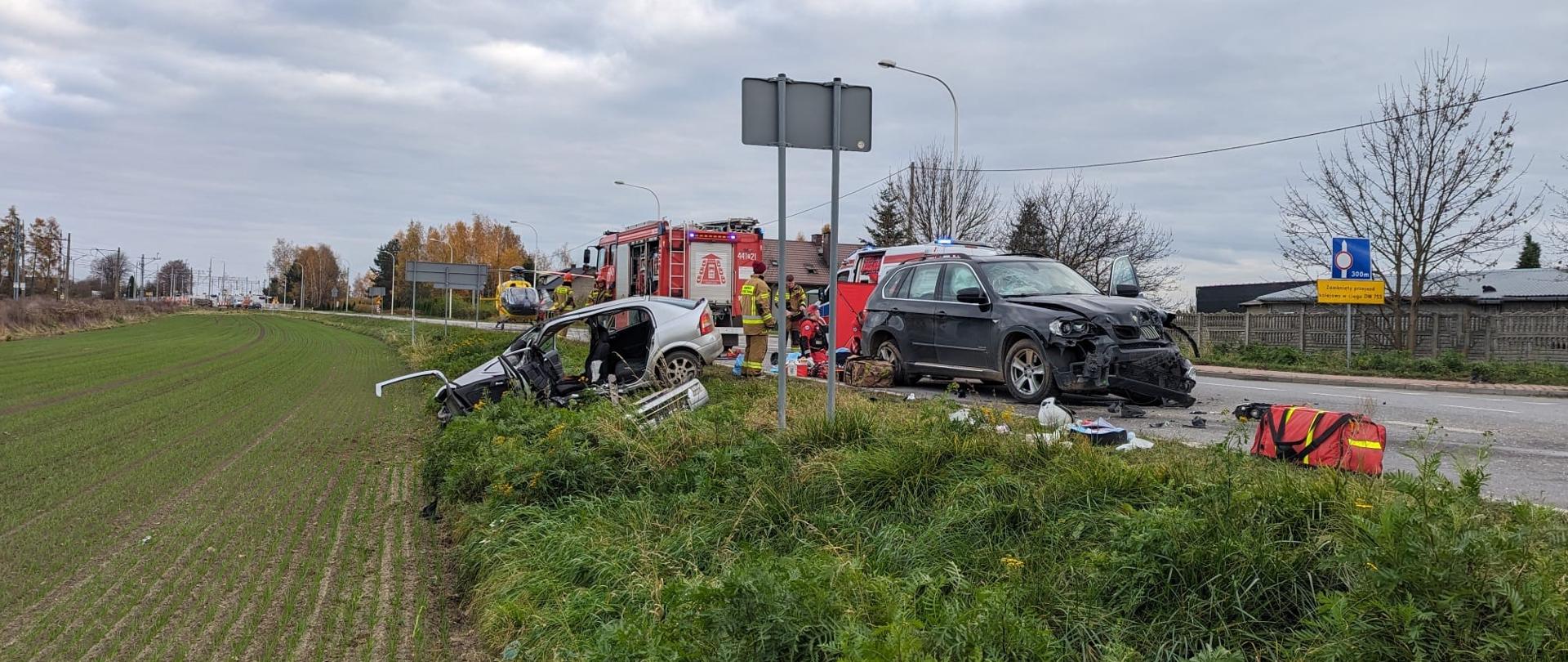 Na zdjęciu wypadek samochodowy (dwa rozbite samochody) w tle strażacy ratownicy medyczni pojazd pożarniczy oraz śmigłowiec LPR.