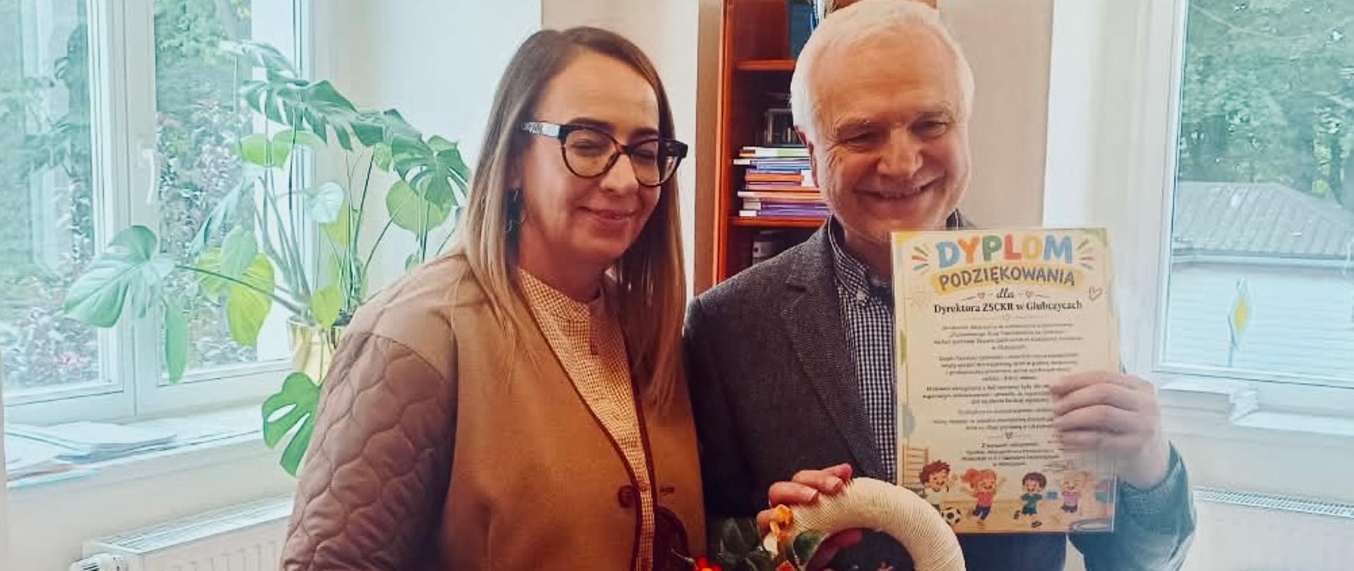 Uśmiechnięta przedstawicielka przedszkola w okularach i brązowej kurtce stoi w gabinecie obok dyrektora szkoły. Dyrektor trzyma kolorowy dyplom podziękowania i jesienny wianek ze sznurka ozdobiony kwiatami i szyszkami.