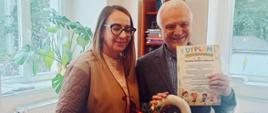 Przedstawicielka przedszkola stoi w gabinecie obok dyrektora szkoły. Dyrektor trzyma kolorowy dyplom i jesienny wianek ze sznurka ozdobiony kwiatami i szyszkami.