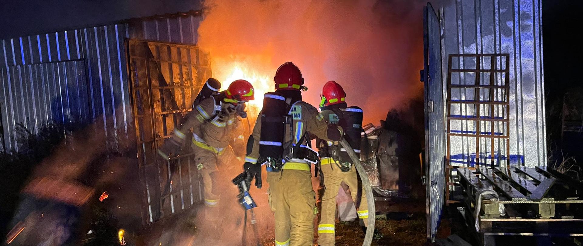 Trzech strażaków w piaskowych ubraniach ochronnych ubranych w sprzęcie ochrony dróg oddechowych wchodzą do środka palącego się magazynu widoczne płomienie oraz dym. Po bokach otwarte metalowe drzwi magazyny z jednej strony kilka palet strażacy trzymają w reku kamerę termowizyjna oraz wąż