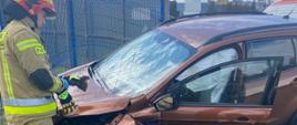 Samochód brązowy z rozbitym przodem przy nim strażak za samochodem ogrodzenie koloru niebieskiego przy otwartych drzwiach samochody od strony kierowcy lezy wyrwane zawieszenie samochodu.