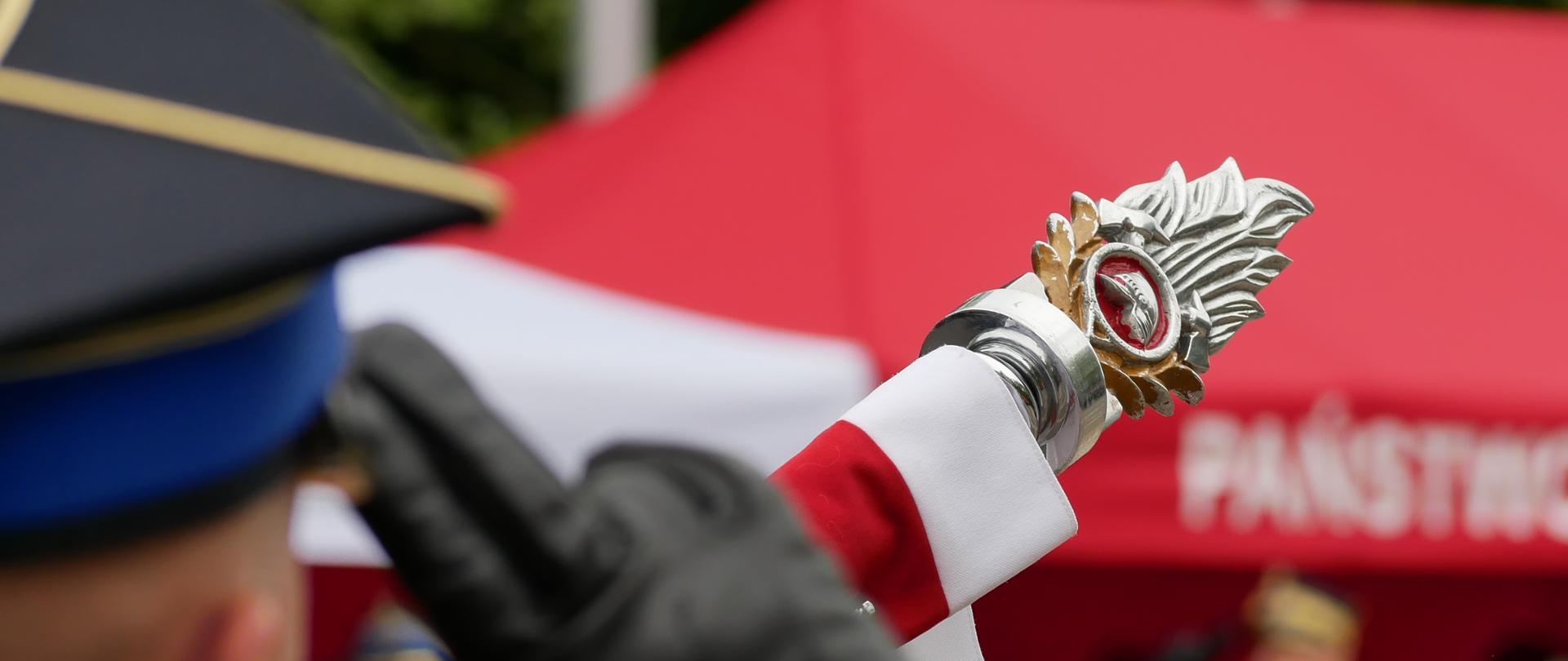 drzewiec sztandaru z biał oczerwoną wstęgą na rozmazanym czerwonym tlenamiotu oraz strażaków