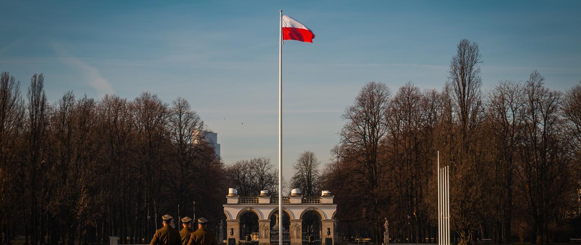 fotografia przedstawia trzech polskich żołnierzy maszerujących po placu, na którym stoi maszt z zawieszoną biało-czerwoną flagą